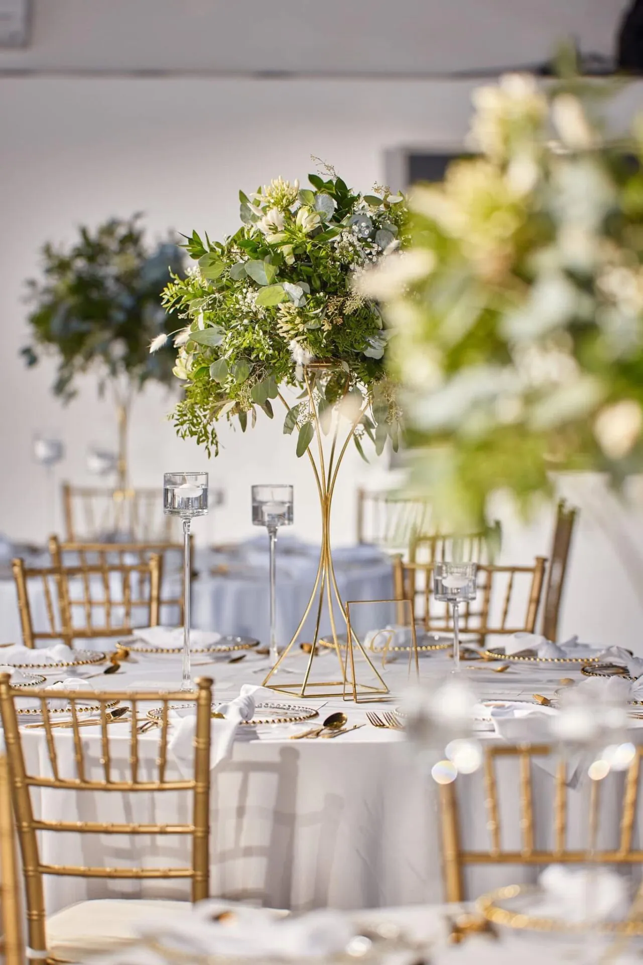 Svadobná výzdoba so zlatými stoličkami a zelenými kvetinovými aranžmánmi v zlatom oblom stojane